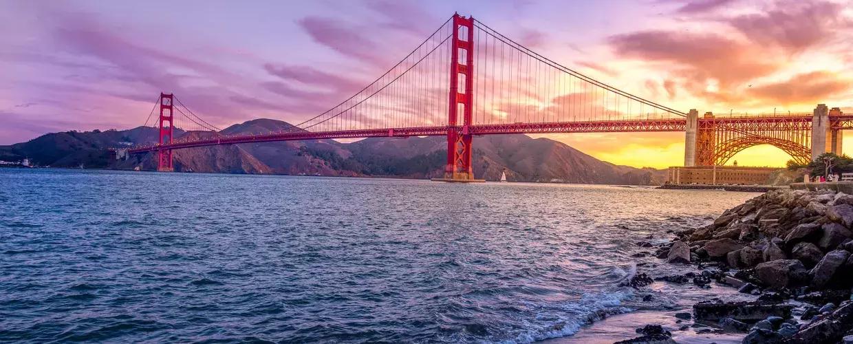 的 金门大桥 at sunset with a multicolored sky 和 the San Francisco Bay in the foreground.